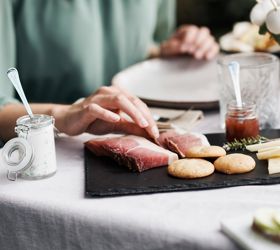 Eine Person greift nach einer Scheibe Speck auf einer Servierplatte mit Südtiroler Spezialitäten wie Käse, Brot, Kräuterquark und Marmelade.
