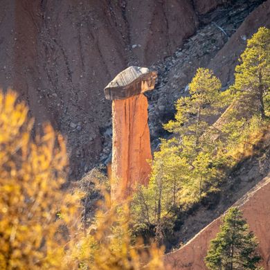 Erdpyramide mit Deckstein inmitten von Bäumen und herbstlich gefärbter Landschaft.