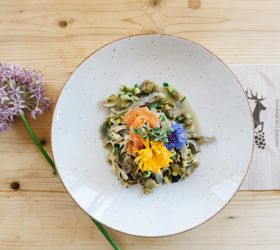 Teller mit Pasta, Gemüse, Lachs und essbaren Blüten, serviert auf einem hellem Holztisch mit einer Blume.