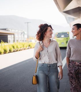 Two women are chatting and smiling at a modern bus stop, with a green bus in the background.